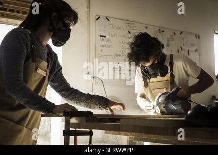 View of a man and a woman wearing protective masks and aprons while using a circular saw to cut a wooden board in a workshop in Lisbon, Lisbon, Portug Stock Photo
