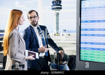 Colleagues on business trip checking gate on screen in busy airport lounge area, waiting for boarding international flight. Man and woman in business attire for corporate travel, Stock Photo