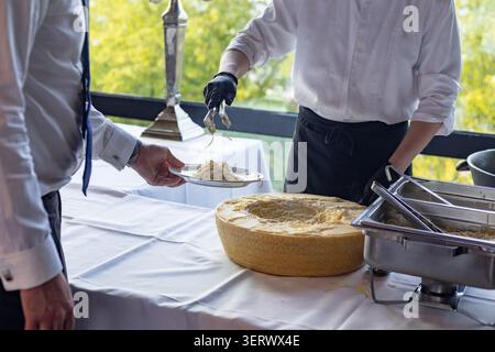 Two people are in a dining area where food is being served. One person holds a plate while another serves pasta from a large cheese wheel to the plate Stock Photo
