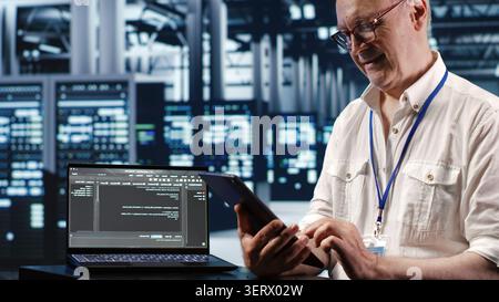 Elderly worker in data center, solving tasks on tablet while running scripts on laptop terminal, certified engineer amidst servers, accessing databases and manipulating lines of code. Stock Photo