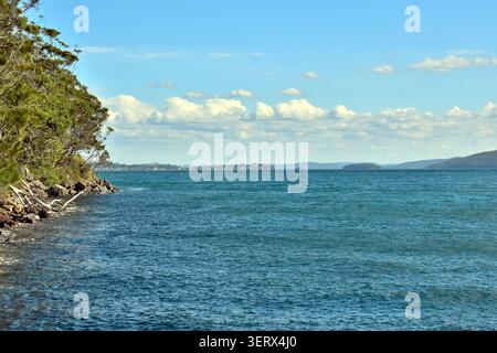 A rocky shoreline lined with dense green trees meets a wide expanse of deep blue water, stretching toward distant land and a small settlement. Stock Photo