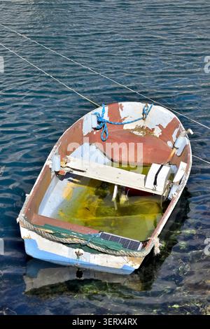 A small, worn rowboat floats gently on rippled water, its chipped paint revealing layers of blue, white and brown. Stagnant yellow water pools inside Stock Photo