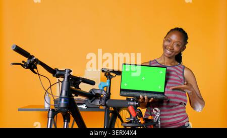 African American woman holds copy space isolated screen laptop, using it to advertise bike repair shop, studio background, mechanic promoting bicycle fixing service with chroma key notebook. Stock Photo