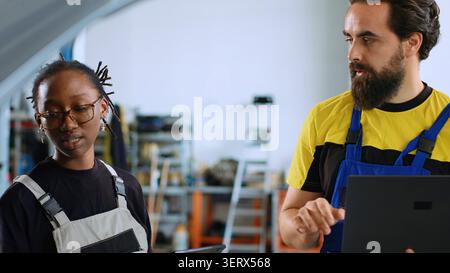 Engineer and colleague doing car maintenance in repair shop using tablet and laptop to keep track of operations, coworkers in garage workplace looking over checklist on devices while fixing vehicle. Stock Photo