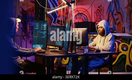 Evil hackers doing data breaches and financial theft in graffiti painted hideout, rogue programmers stealing users bank account credentials, targeting low security passwords. Stock Photo