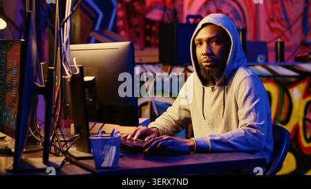 Portrait of African American hacker in abandoned warehouse using phishing technique that tricks users into revealing sensitive information, hooded cybercriminal stealing passwords. Stock Photo