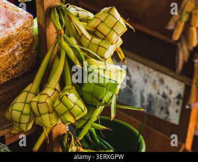 A bunch of fresh, green woven young coconut palm leaves shaped into ketupat pouches, hanging from a wooden structure at a traditional market stall. Stock Photo