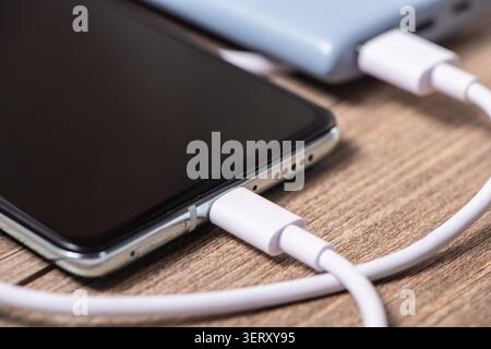 Smartphone connected to a power bank, receiving charge via a white usb-c cable on wood Stock Photo