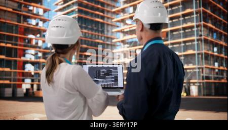 Two Architects And Engineer Discuss 3D BIM Model On Busy Construction Site. Stock Photo
