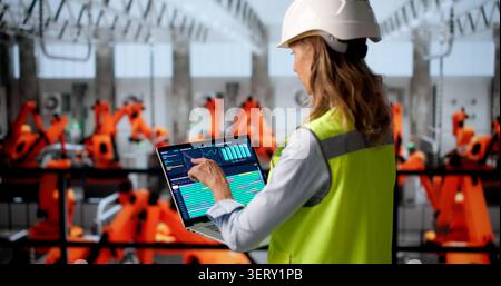 Mature Woman Engineer Designs Digital Systems Using Laptop In Busy Factory. Stock Photo