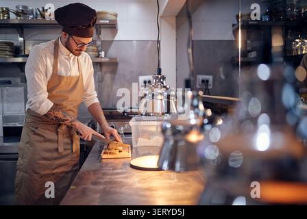 chef slicing bread with knife on wooden board. . Cook in Professional Modern  kitchen, restaurant concept Stock Photo