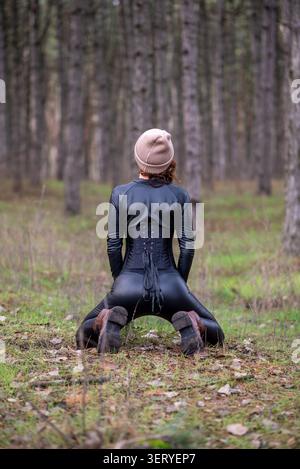 Rear view of a woman wearing a black leather corset, leggings and knit beanie, kneeling on the forest floor among pines for an outdoor fashion portrai Stock Photo