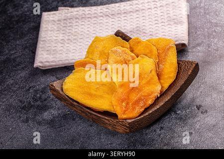 Yellow dried mango slices arranged on dark wooden plate with beige napkin on gray stone background. Healthy tropical fruit snack, dehydrated food phot Stock Photo