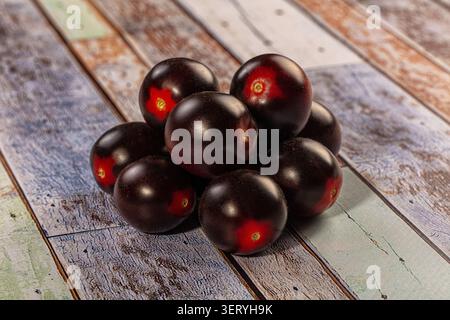 Pile of fresh dark cherry tomatoes with deep red accents rests on weathered wooden planks. Natural light highlights glossy skin, creating healthy, rus Stock Photo