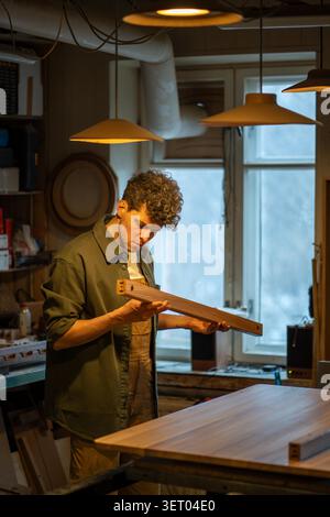 Serious focused man carpenter working. Concentrated male pro master joiner at workplace. Interested guy professional woodworker preparing wooden plank, polishes removes dust with towel napkin cloth. Stock Photo