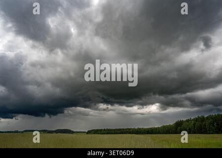 Large dark storm clouds dominate the sky above a rural meadow and forest landscape Stock Photo