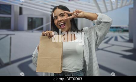 Woman holds brown paper bag while making a peace sign at a building entrance, smiling with teeth visible; playful confidence. Stock Photo