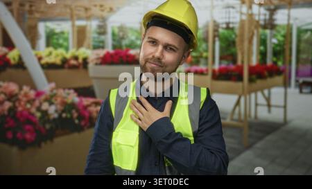 Man construction worker with hand on chest wearing yellow hardhat and high visibility vest amid flower shop stalls in building; gratitude. Stock Photo