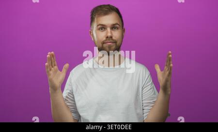 Man, bearded and smiling, shows open hands and palms in magenta studio wearing white t shirt with relaxed posture and direct eye contact; friendly ope Stock Photo