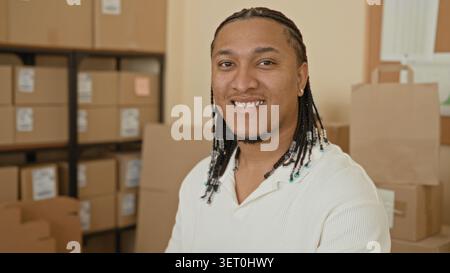Man smiling beside stacked cardboard parcels and boxes in a building packing area; small business optimism. Stock Photo