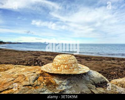 A straw hat sits on a rock at the beach. The sea is visible in the background along with some clouds. The sun shines brightly on the scene. Stock Photo