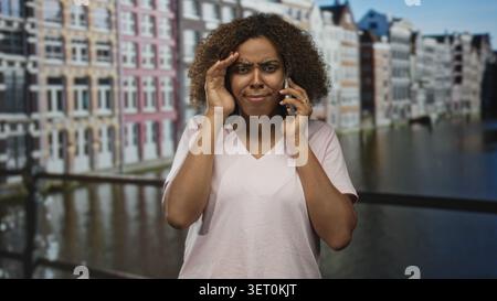 Woman on a canal bridge holding smartphone to ear with hand on forehead in front of amsterdam building; worry phone problem. Stock Photo