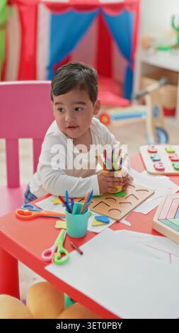 Boy with hand holding colored pencils and wooden shape puzzle on red table in building; curious concentration. Stock Photo