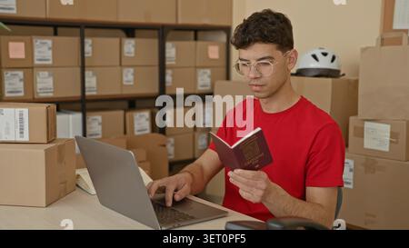 Young man in red tshirt holds czech passport and types on laptop while checking packages and boxes in building; entrepreneurship planning focus. Stock Photo