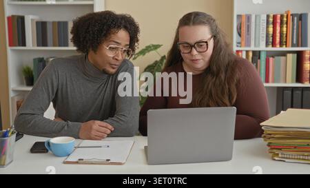 Man and woman leaning over laptop at desk with clipboard, coffee cup and stacked folders, man making a fist gesture while studying the screen in a bui Stock Photo