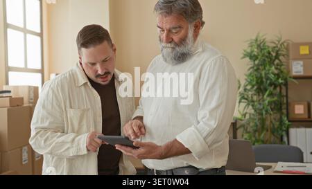 Two men point finger to tablet while checking stacked packages and discussing parcel labels in a building small business delivery office; collaboratio Stock Photo