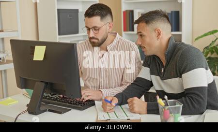Two men at computer, one typing on a keyboard and the other pointing to a clipboard while reviewing a form in an office building; concentration teamwo Stock Photo