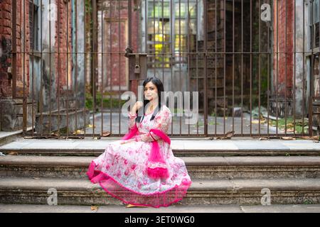 A woman in a traditional floral-patterned dress with pink accents poses on a decorative stone staircase in front of an old brick building with metal gates. Stock Photo