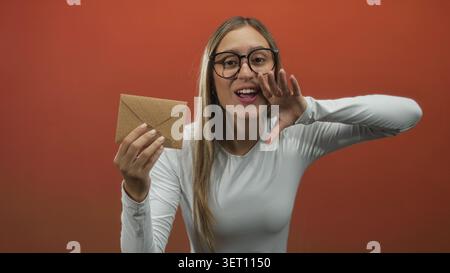 Young woman holding kraft envelope with outstretched hand and smiling gesture in studio; joy surprise greeting. Stock Photo