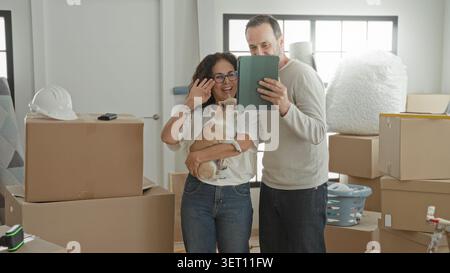 Couple holding small dog and tablet together while standing among packed moving boxes in a new house; new beginnings hopeful. Stock Photo