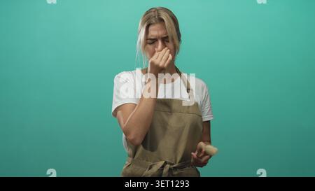Woman makeup artist young blonde wearing apron pinches nose while holding brush in studio with teal backdrop; disgust reaction. Stock Photo