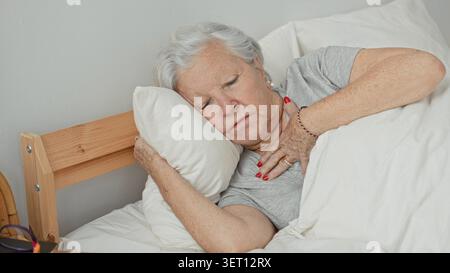 Woman clutching chest with hand while reclining on pillow in bed inside a building, red fingernails visible, eyes closed; pain vulnerability. Stock Photo