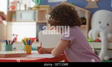 Girl hispanic child drawing with colored pencil at small red table in daycare building, leaning over paper surrounded by toys and crayons; calm concen Stock Photo
