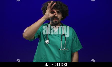 Man doctor in green scrubs with stethoscope making ok sign with hand over eye in studio with deep blue background; playful curiosity. Stock Photo