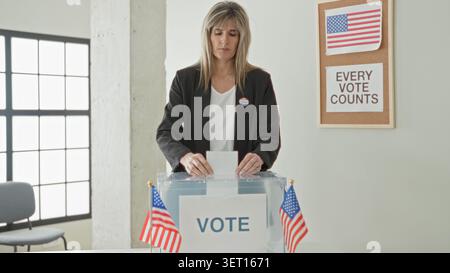 Woman inserting ballot into transparent ballot box with sticker visible in polling building interior, american flags and 'every vote counts' sign; civ Stock Photo