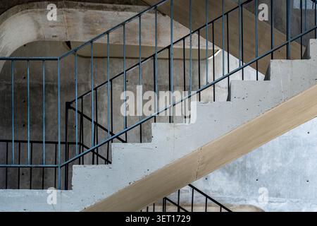 An architectural photograph highlighting a brutalist raw concrete zig-zag staircase. Strong daylight cuts through the scene, casting high-contrast, dr Stock Photo