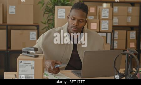 Man holds smartphone to ear and checks laptop among stacked parcels and barcode scanner on desk in a building; small business success joy. Stock Photo