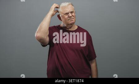 Senior man in burgundy t shirt scratching head with right hand while wearing glasses in studio grey wall setting; confusion reflection pondering. Stock Photo