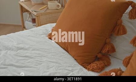 Woman adjusting a textured cream throw pillow on a queen bed in building bedroom beside a rattan nightstand with an orange knit blanket; calm morning Stock Photo