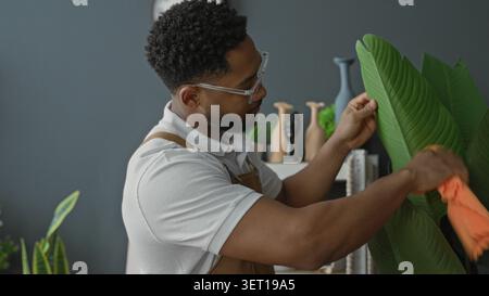 Man florist in brown apron wiping large tropical plant leaves with bright orange cloth inside studio workspace, tending foliage and dust; focused care Stock Photo