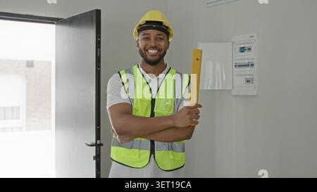 Man in yellow hardhat and hi vis vest holds yellow measuring ruler with crossed arms and smiles inside building; professional confidence. Stock Photo