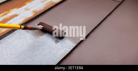 painting tin metal roof with roller and brown paint. rusty house roofing restoration Stock Photo