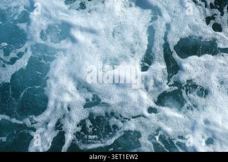 Ocean waves crashing against rocks creating foam and bubbles in clear blue water under bright sunlight Stock Photo