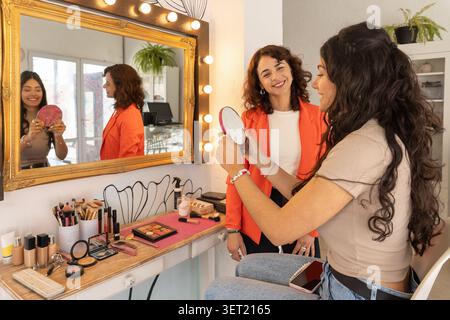 Satisfied client smiles while checking her completed makeup look in a hand mirror as the makeup artist stands beside her in a beauty salon. Stock Photo