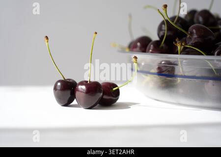 Fresh dark sweet cherries with stems beside a clear plastic bowl on a clean white background Stock Photo