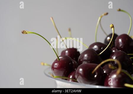 Fresh dark sweet cherries with stems beside a clear plastic bowl on a clean white background Stock Photo